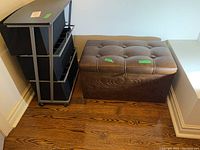 Metal frame storage cabinet with three cloth storage bins and brown leather tufted ottoman side by side on wooden floor.