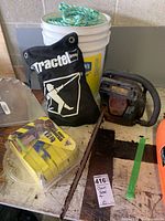 Photo showing gas powered chain saw, full body harnesses, nylon rope in bucket, and tied yellow straps in plastic container on wooden table.