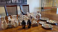 Photo showing assortment of decorative china plates, vases, figurines, and serving bowl arranged on a wooden table with chairs in background