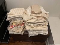 Pile of various folded cream and off-white lace doilies, cloths, table runners, and napkins stacked on a wooden chest.