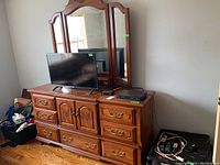 Front angled view of dresser and attached triple mirror with items on top including a TV and remote.