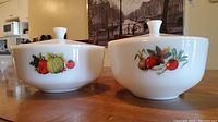 Two white vintage F Brand glass serving bowls with vegetable prints on the side, both with lids, one lid has a chip on top.