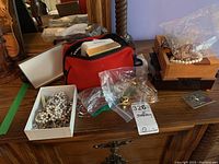 Overview of the jewelry lot showing red bag, plastic bags with assorted jewelry, a box with pearl necklaces, and small wooden jewelry boxes with additional jewelry on top