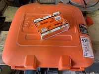 Closed orange plastic hard carrying case with two boxes of Paslode 16GA nails on top. The case has Paslode branding.