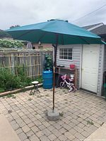 Patio umbrella standing upright on a paved patio, showing umbrella fully open with green canopy and wooden pole in a concrete base, outdoor setting with garden and house in background.