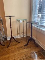 Three plant stands: two dark wooden round top stands with tripod legs and one metal frame stand with glass top; scratches visible on wood.