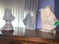 Three religious statues on wooden surface by window; one male bust on pedestal, one figurine of man, woman and child, one female bust on marble base.
