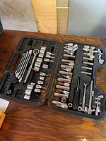 Open black plastic carry case displaying various chrome sockets, ratchet wrench, wrenches, and extensions.