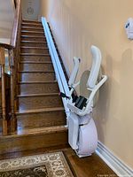 Savaria K2 stairlift installed at bottom of wooden staircase with white metal rail divided into two pieces, showing the entire rail and stairlift unit.
