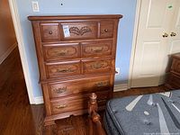 Front view of wooden tall boy dresser featuring five drawers with carved floral detail on center top drawer. Mixed hardware with round knobs and brass handles.