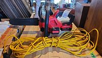 Photo shows red and black Craftsman cordless chainsaw with battery installed and yellow extension cord coiled and lying on the table