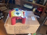 Yellow and red plastic decorative wall clock resting on top of a cardboard box with original boxed cuckoo clock next to it.