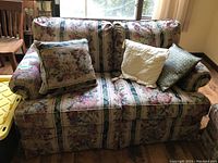 Full view of the floral patterned fabric loveseat with three decorative pillows on it. The loveseat has rolled arms and two main cushions.