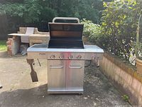 Full front view of stainless steel grill showing hood open, side shelves, cabinet base and spatulas hanging on left shelf