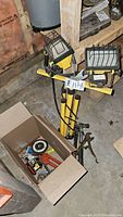 Two yellow Noma work lights with black protective cages, standing with tripods. An open box containing various small tools and hardware measuring tapes, screwdrivers, tape rolls.