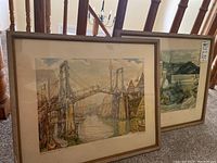 Photo showing two framed signed prints leaning against a stair railing, one with a bridge scene and the other with a snowy church scene.