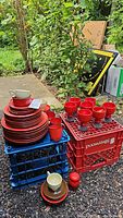 Stack of red Thomas Flame plates, cups, saucers, and red pottery goblets arranged on crates outdoors with some cardboard boxes and greenery in background.