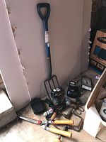 Pitch fork with blue handle, vintage hand crank drills, drill bits, large pruners, Craftsman DieHard drill and extra battery shown on concrete floor near a wall.