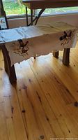 Wooden bench covered with embroidered white fabric featuring roosters and chicks by window on wooden floor.