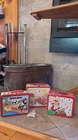 Set of three vintage metal lunchboxes sitting on stone surface near fireplace, showing red handles, colorful front art, and signs of wear