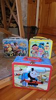 Three metal lunchboxes in a group, showing front artwork from Fraggle Rock, Sesame Street, and Thomas the Tank Engine.