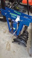 Blue steel pipe bender frame with hydraulic cylinder and lever, seven bending dies on floor