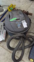 Top view of black and gray cylindrical shop vacuum showing 6.5 gallon capacity label, attached vacuum hose coiled on the front, and some surrounding garage items.