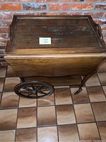 Full view of vintage wood rolling trolley with drop leaves and wheels against brick background on tile floor.