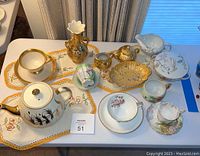 Porcelain pieces and doilies on a table showing cups, teapot, decorative bowls, creamer, sugar bowl, and embroidered doilies