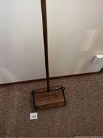 Full view of antique wooden and metal floor sweeper with long handle standing upright against wall on carpet.