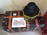 Full view of the large vintage steamer trunk with orange bands, the framed watercolor print, and the black wool bowler hat resting on the Dobbs hatbox.