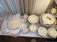 Overview of white bone china plates with gold leaf trim, cups with saucers, covered casserole dish, silver-plated basket, and oval serving bowl arranged on table.