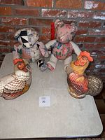 Group photo showing two handmade stuffed bears and two ceramic chicken figurines arranged on a table against a brick wall.