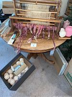 Wooden loom setup with hanging warp threads in pink and white, on a wood table with yarn spools nearby.