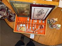 Full lot image showing silver plate flatware set in orange case, two framed artworks behind, Victorian cranberry glass compote on right, figurines in background
