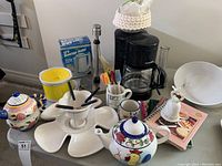 Photo showing coffee maker, ceramic teapot, mini slow cooker, divided serving dish, and assorted kitchen mugs and utensils on table.
