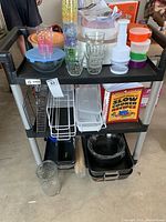 Image of black rolling utility cart with three shelves holding kitchenware including glassware, plastic containers with lids, roasting pans, mixing bowls and chopping tool.