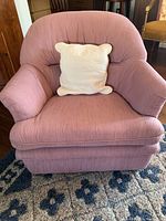 Soft pink cushioned armchair with cream colored throw pillow on top, sitting on the patterned area rug