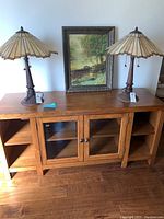 Full view of wooden cabinet with two glass doors in center, open wooden shelves on sides, photographed with two lamps and a framed painting on top.