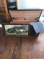 Open solid wood hope chest with cedar interior visible and black metal hardware, placed on a wood floor. Western-themed framed painting with horses and riders leaning against the chest.