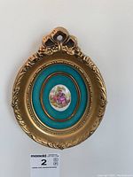 Gold-colored oval frame with a turquoise background porcelain miniature plaque showing a woman holding a guitar with a man and dog, decorative floral bow top, approx 5-7 inches tall.