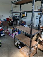 Full view of metal shelving unit with four wooden shelves in a garage setting. Shelves hold various items but items are not included in lot.