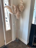Full view of pampas grass stalks arranged in tall clear glass vase placed on floor near door and wall corner.