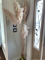 Five beige pampas grass stems arranged inside a tall clear cylindrical glass vase near a door with decorative glass window.