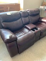 Three-seater brown leather or leather-like electric recliner sofa with padded armrests and built-in cup holders in the center armrest.