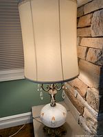 The entire lamp with cream fabric shade illuminated, showing overall height and round shade shape near stone wall.