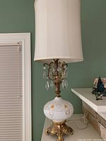 Full view of vintage French inspired table lamp with off-white cylindrical fabric shade, glass body decorated with orange and gold patterns, and brass claw foot base with crystal drops hanging near the socket.