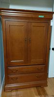 Full front view of wood armoire showing two paneled doors and two drawers below with metal handles, standing on a hardwood floor against a wall.