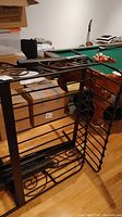Metal single bed frame with decorative scrollwork, showing headboard, footboard, and rails leaning vertically in a room with wooden floor and pool table in background.