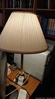 Photo of a 27-inch brass table lamp with beige pleated lampshade on a wooden desk next to books and electronic equipment.